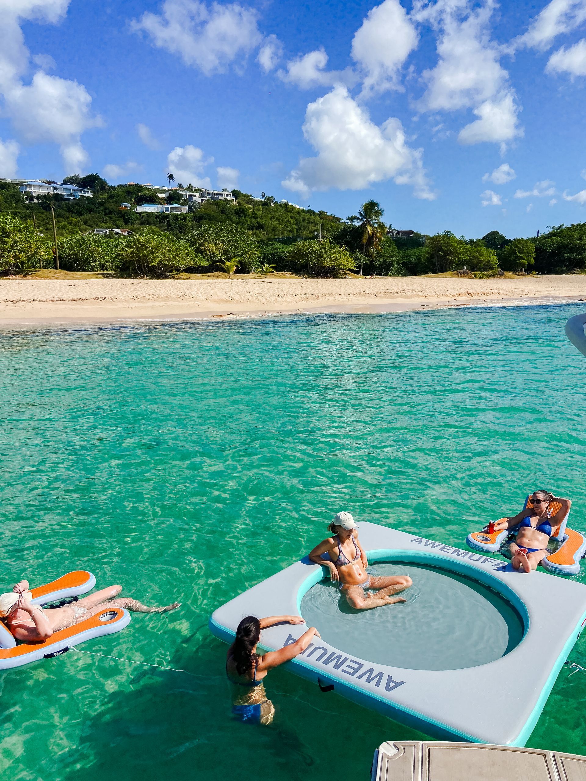 People relax on floating mats and a water trampoline in turquoise water near a beach.