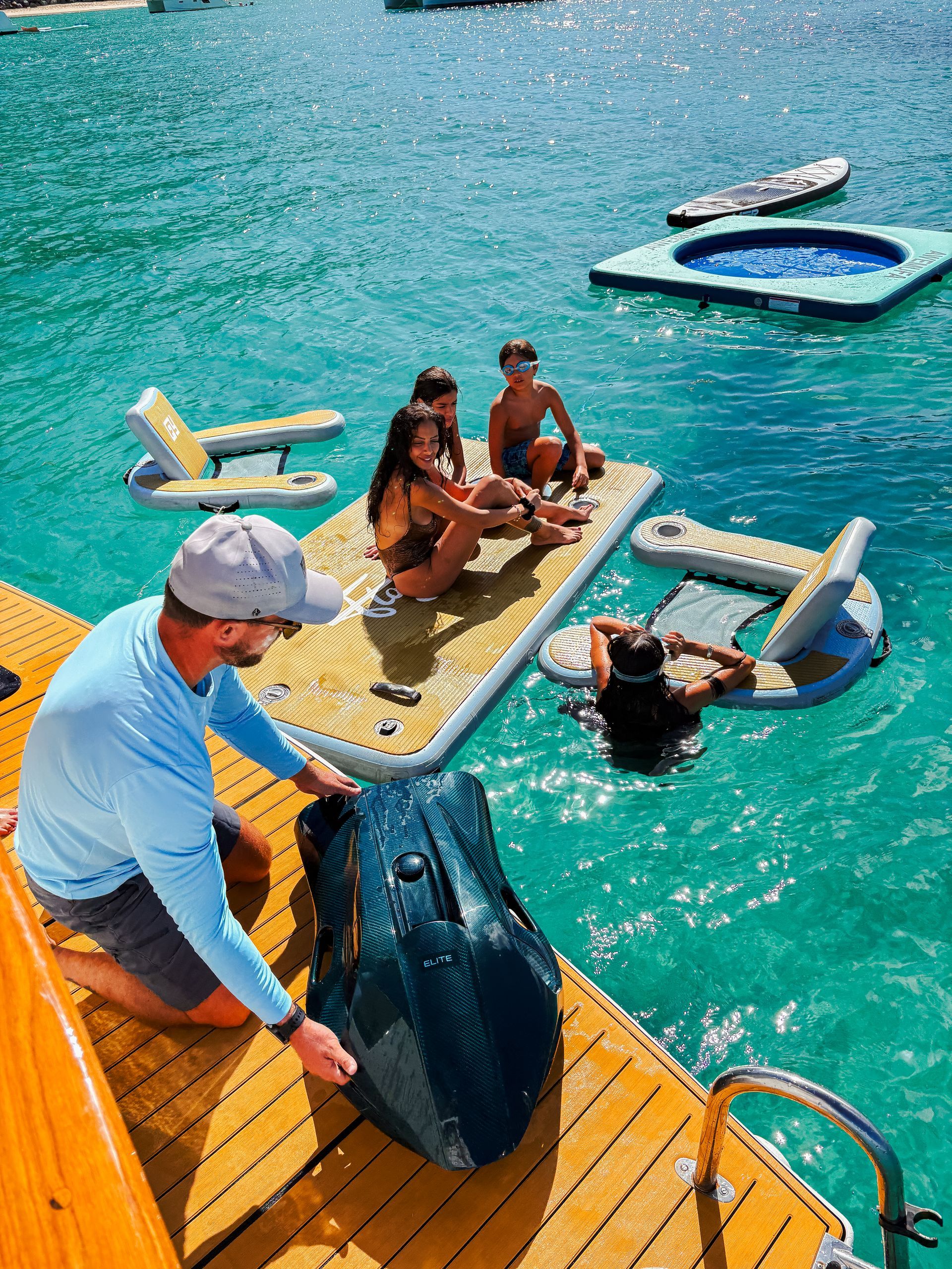 Man on dock with wakeboard, looking at people on water platforms with lounge chairs in turquoise water.