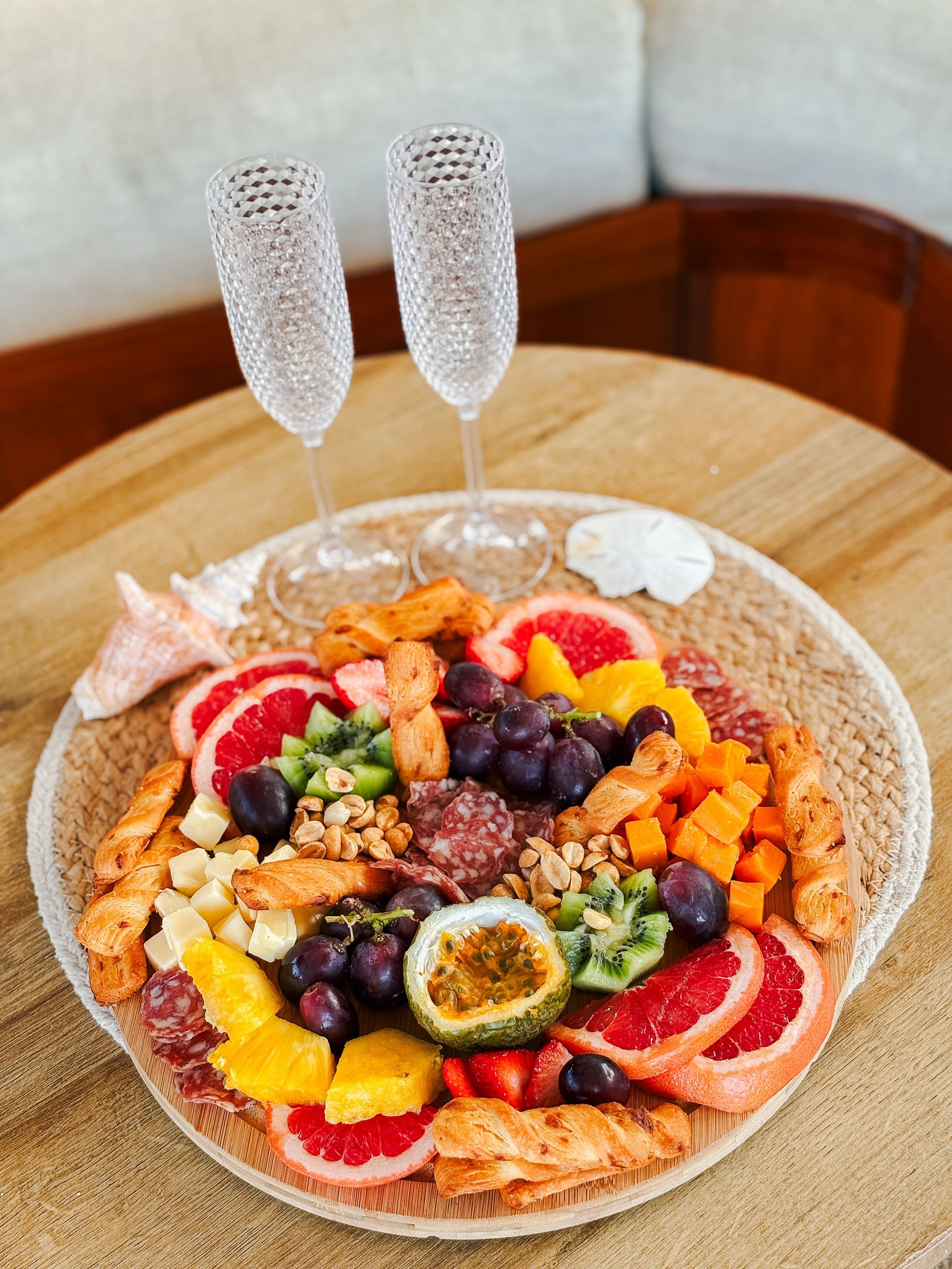 Charcuterie board with fruit, crackers, and meat, paired with champagne flutes on a woven mat.