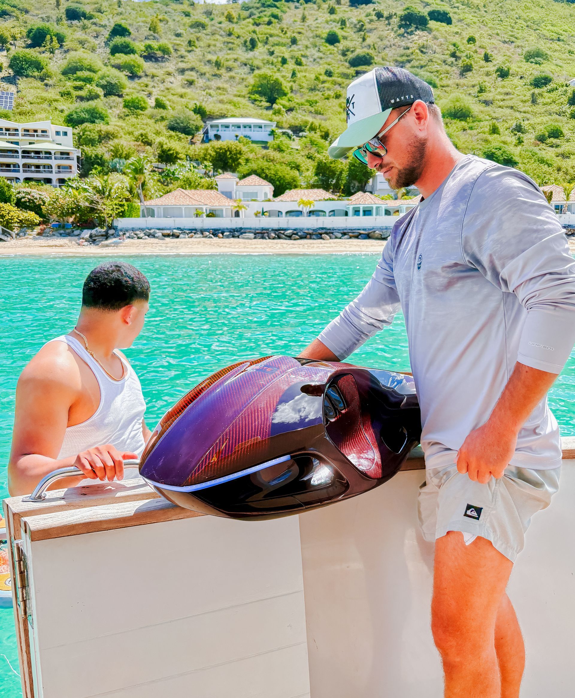 Two men on a boat, one holding a large purple object, near a turquoise ocean and green hills.