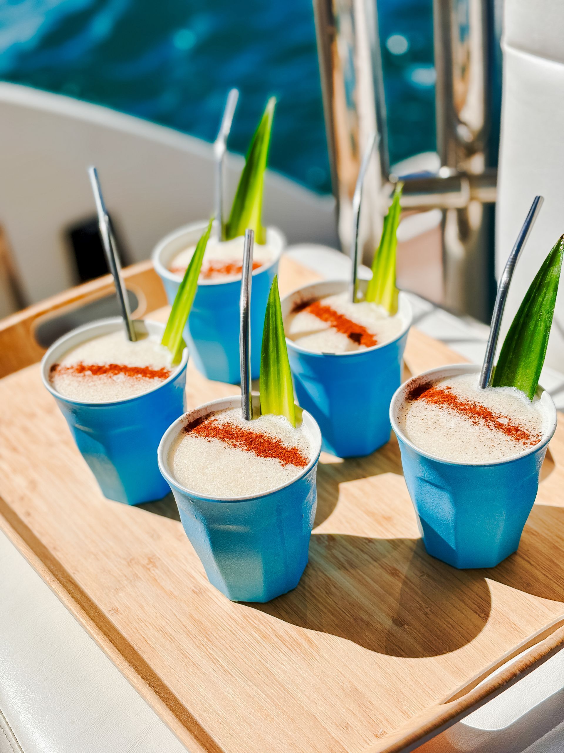 Six blue cups of drink on a wooden tray, each with a celery stick, metal straw, and paprika.
