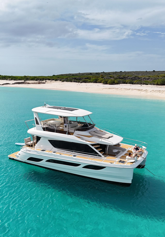 White catamaran in turquoise water near a beach. People are near the boat with what appear to be paddleboards.