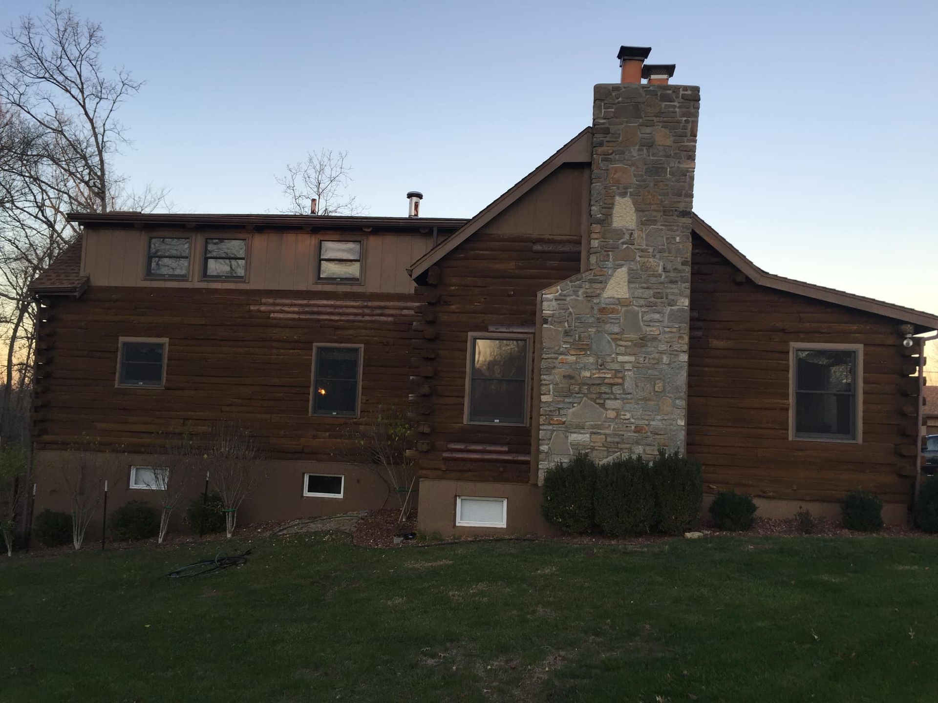 Brown log cabin house with stone chimney, windows, and green lawn.