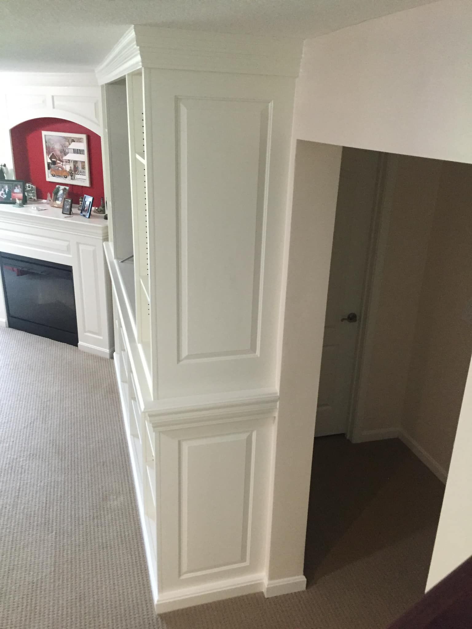 White built-in bookcase and pillar separating a living area with a fireplace from a hallway leading to a door.