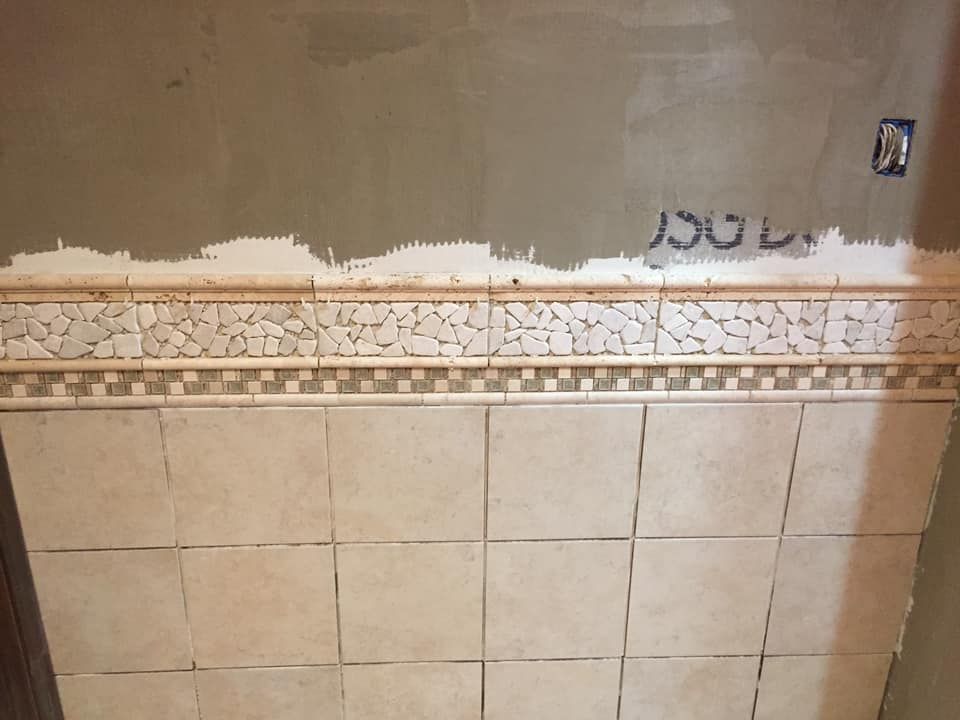 Bathroom wall in progress: beige tiles, mosaic border, unfinished plaster above.