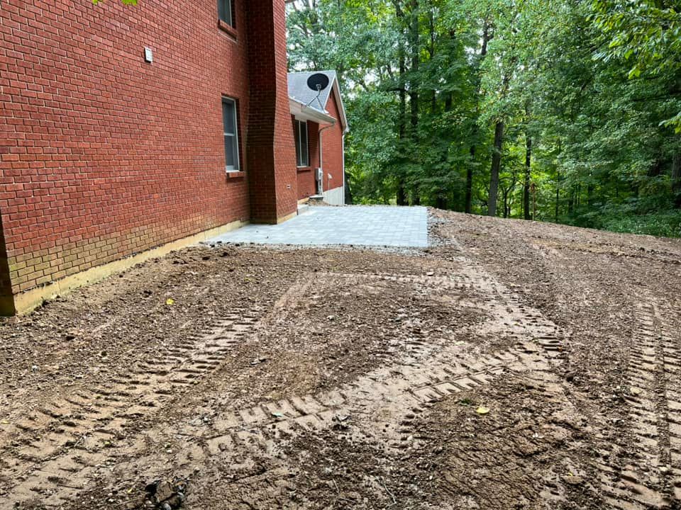 Backyard area with a new gravel patio and graded soil; red brick house and trees in the background.