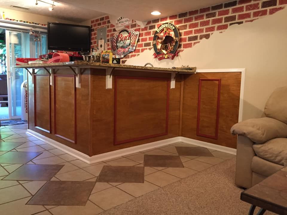 Basement bar with wood paneling, tile floor, and brick accent wall, with a TV, bar signs, and a tan sofa.