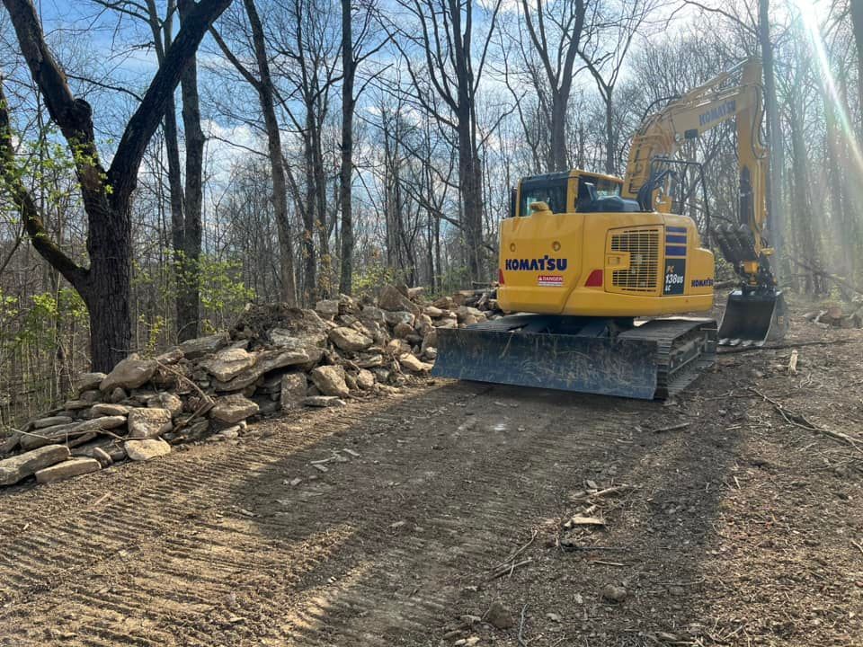 Yellow Komatsu excavator on a dirt path next to a pile of stones in a wooded area.