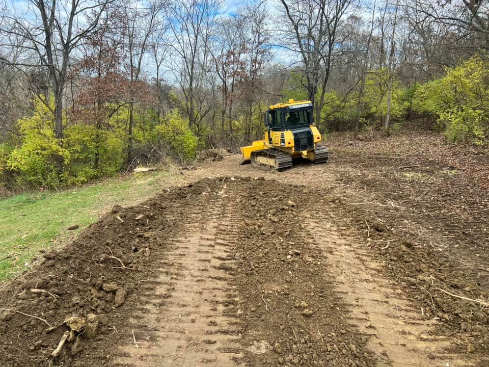 Yellow bulldozer grading a dirt path on a hillside with trees in the background.