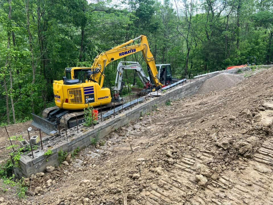Yellow excavator and smaller white excavator on a construction site, near a concrete structure and a hillside.