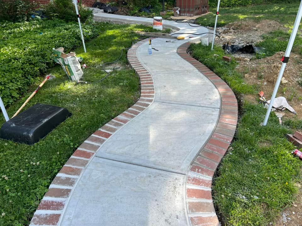 Newly constructed winding concrete walkway with brick border, surrounded by grass and greenery.
