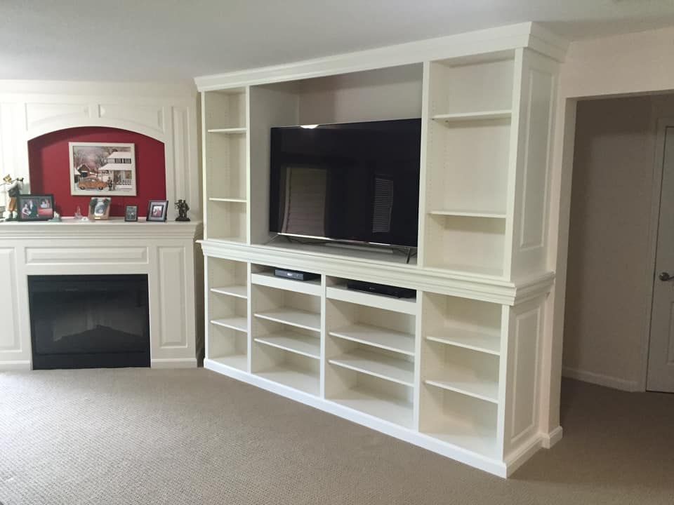 Built-in white entertainment center with shelves, TV, and fireplace in a room with beige carpet.