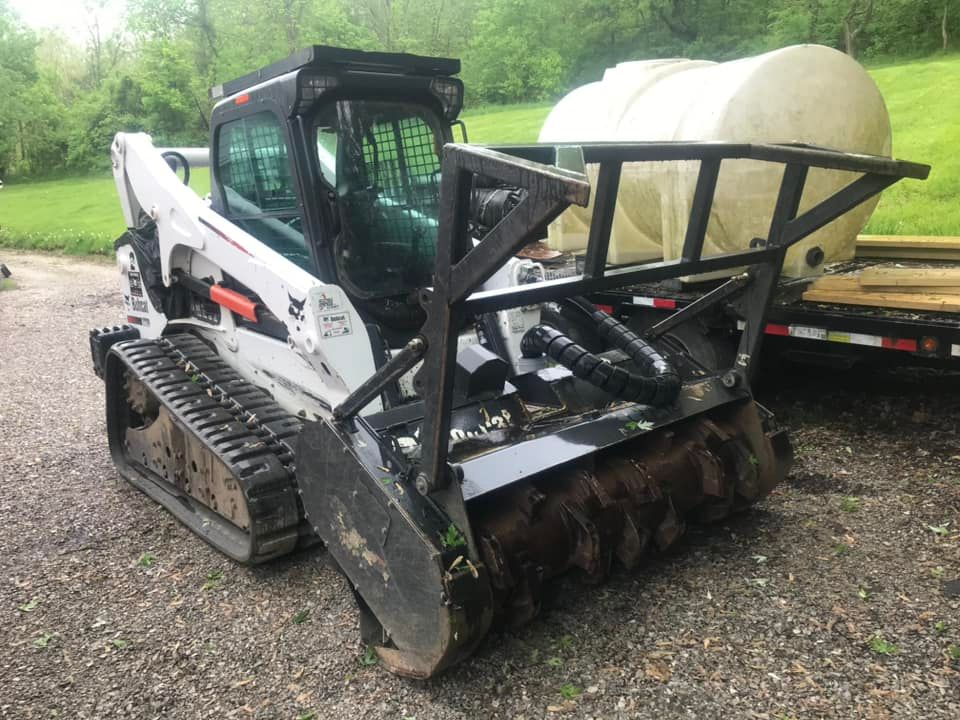 Bobcat track loader with forestry mulcher attachment, cream-colored water tank in background.