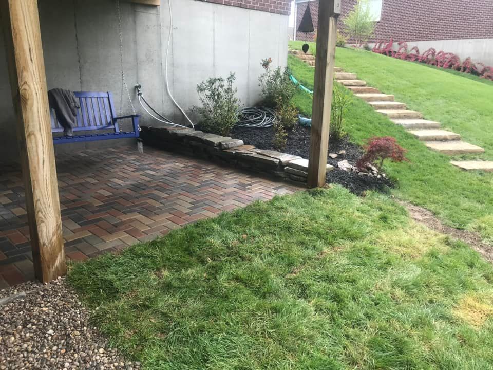 Brick patio with wooden posts, stone wall, blue swing, and a grassy hillside with stone steps.