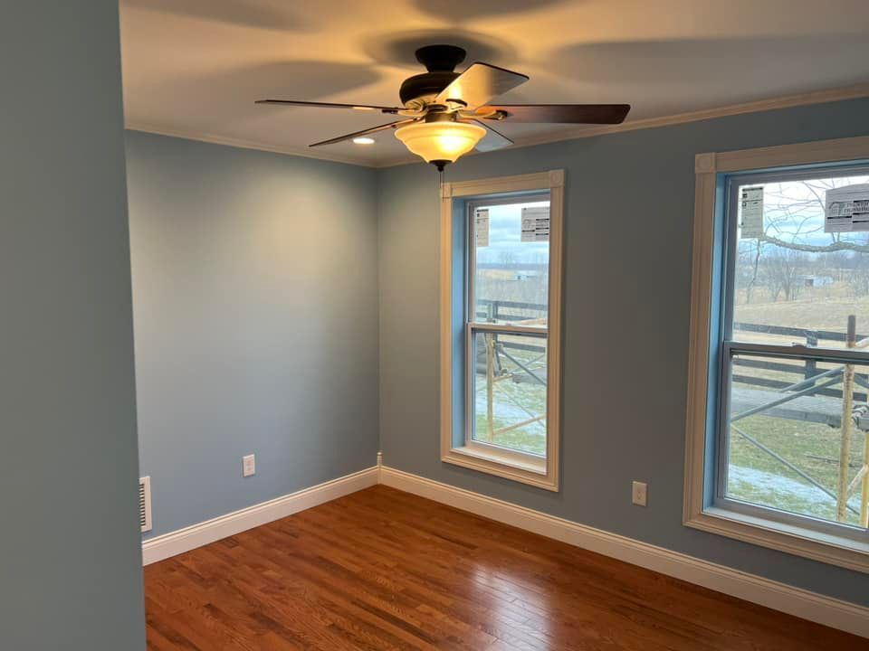 Empty room with blue walls, two windows, wood floor, ceiling fan, and white trim.