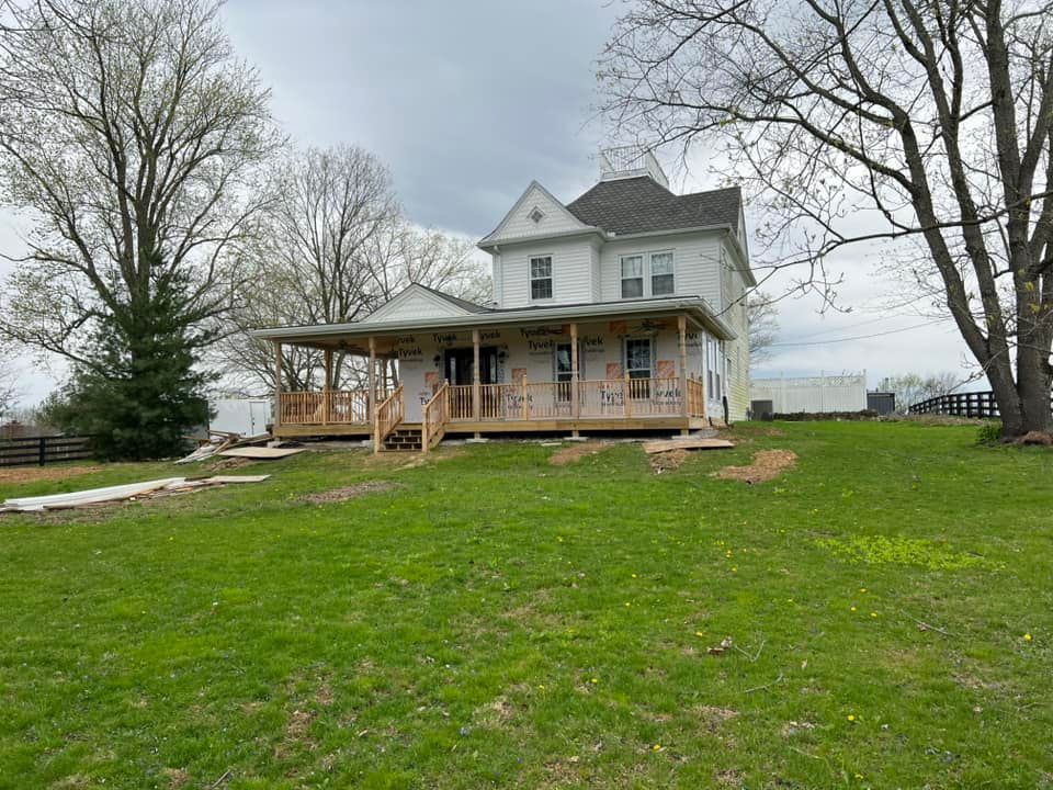 Two-story white house with wraparound porch on a grassy hill; construction in progress. Overcast sky.