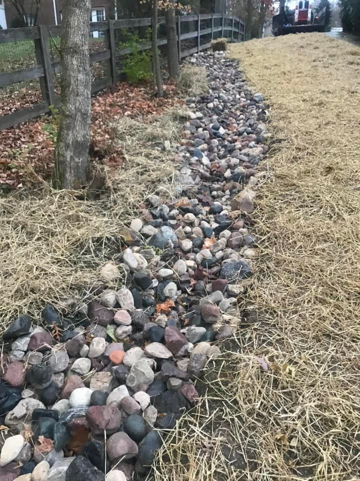 Rocks in a narrow bed alongside a grassy embankment, beside a wooden fence and trees; car in background.