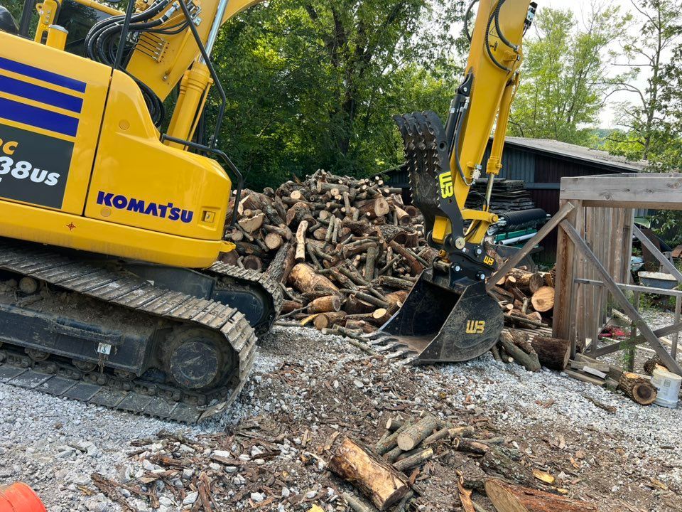 Yellow Komatsu excavator loading firewood on gravel ground.