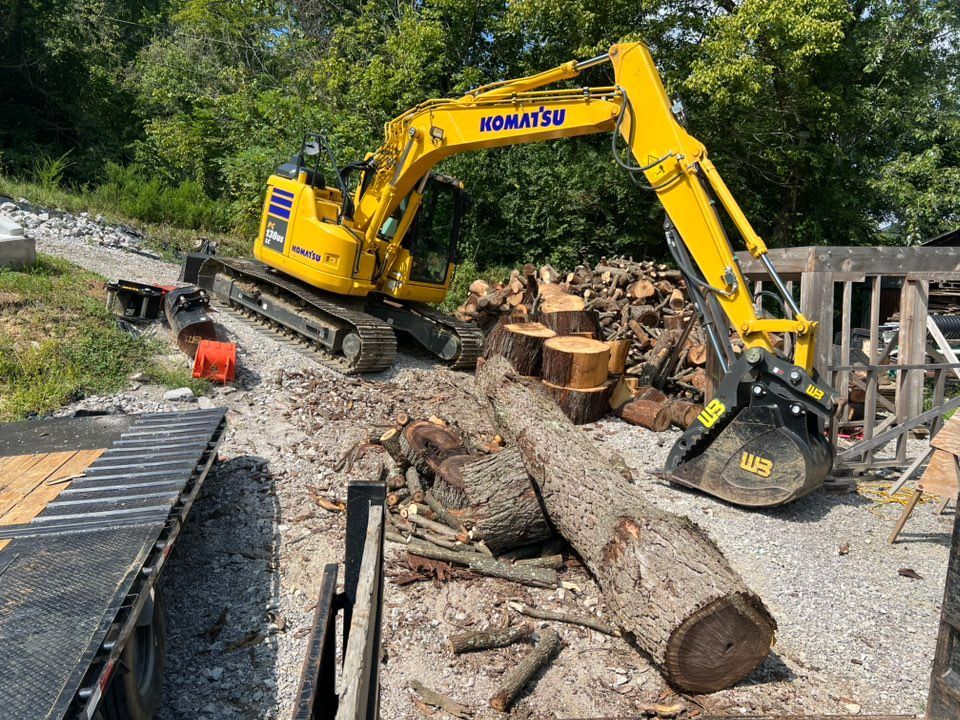Yellow excavator lifting a large log next to a woodpile and trailer outdoors.