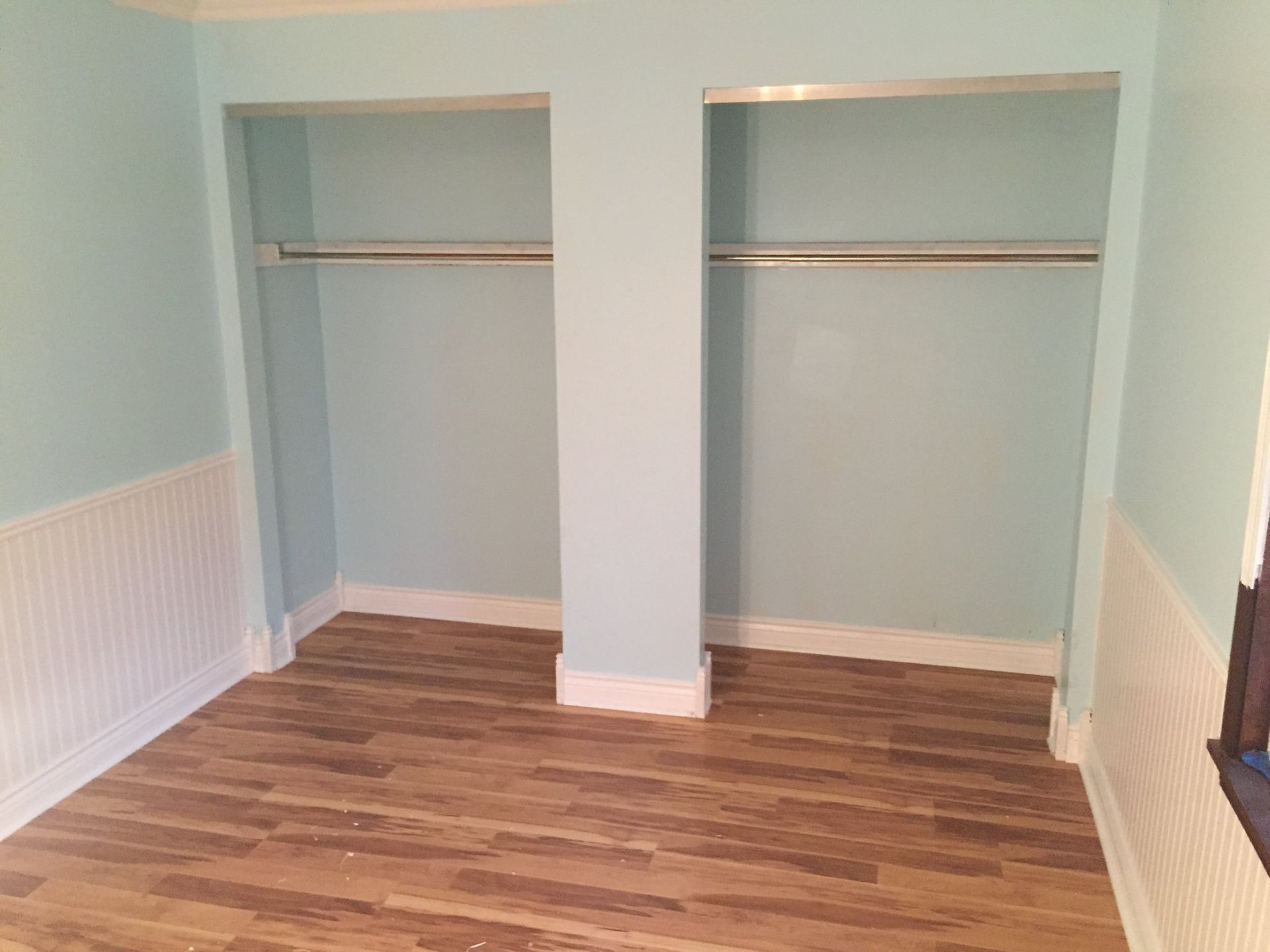 Empty room with wood-look floor, pale blue walls, two closet openings with hanging rods, and white wainscoting.