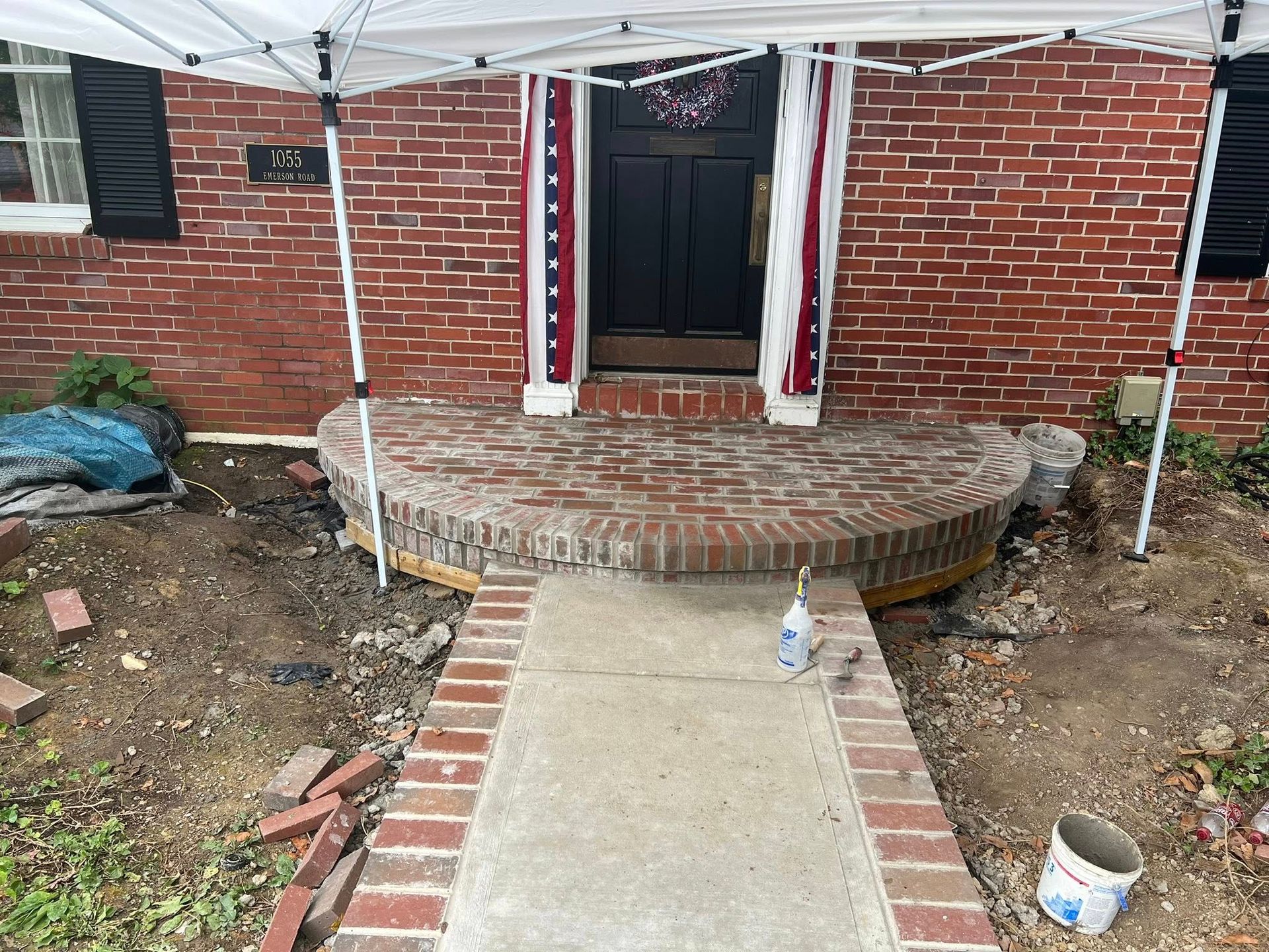 Brick front porch under construction, sidewalk with brick edges, red brick house with tent.