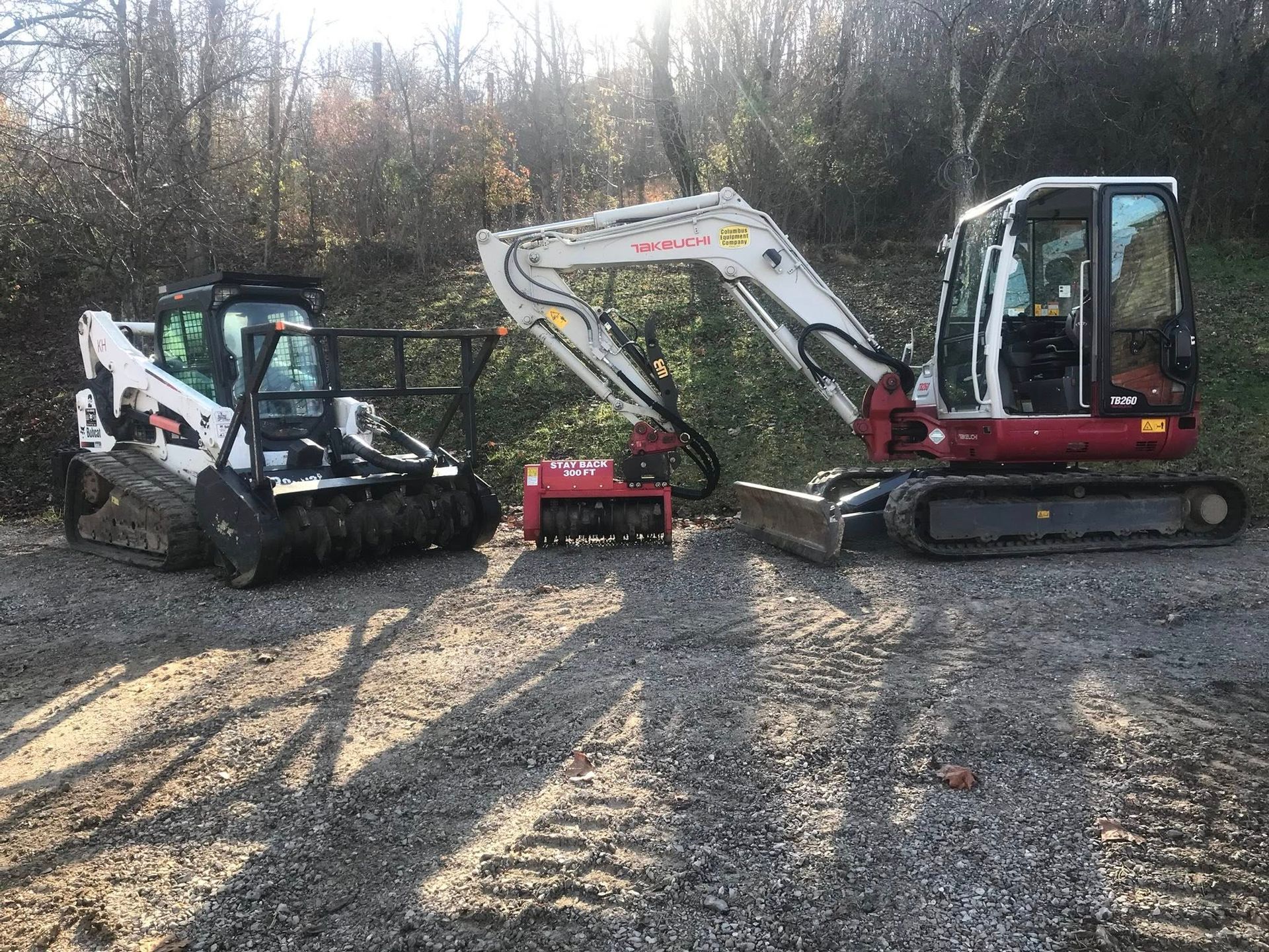 A Bobcat, stump grinder, and excavator on a gravel road next to a wooded area.