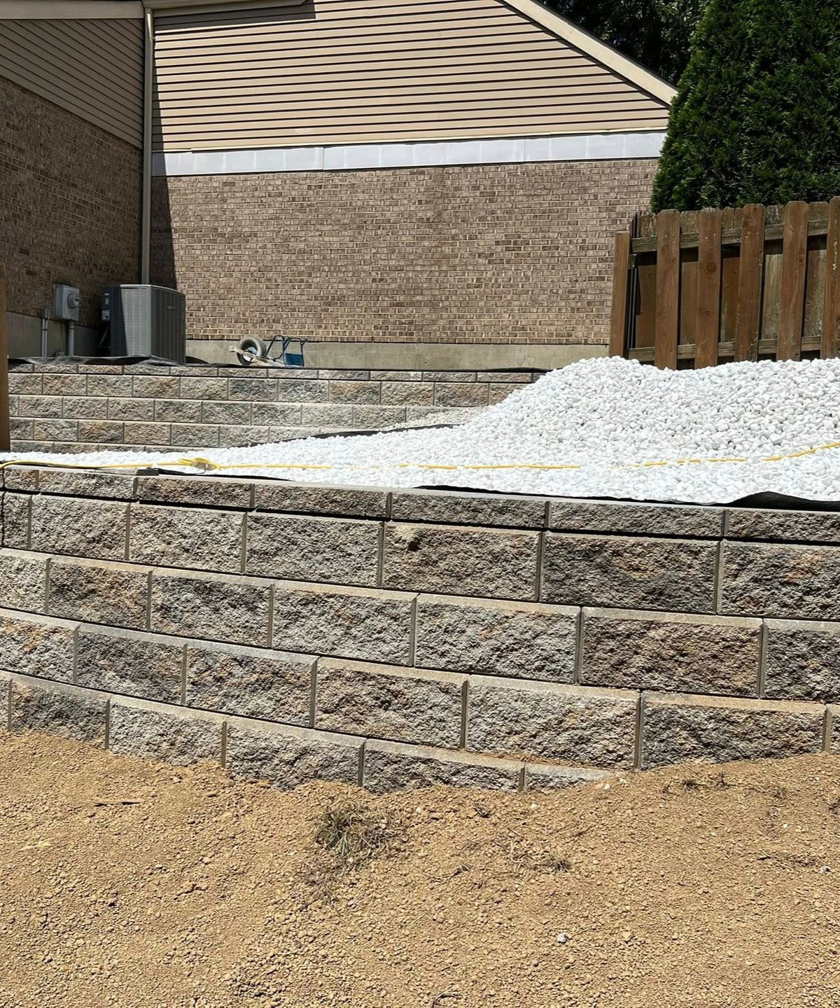 Stone retaining wall in front of a building with gravel and a wooden fence in the background.