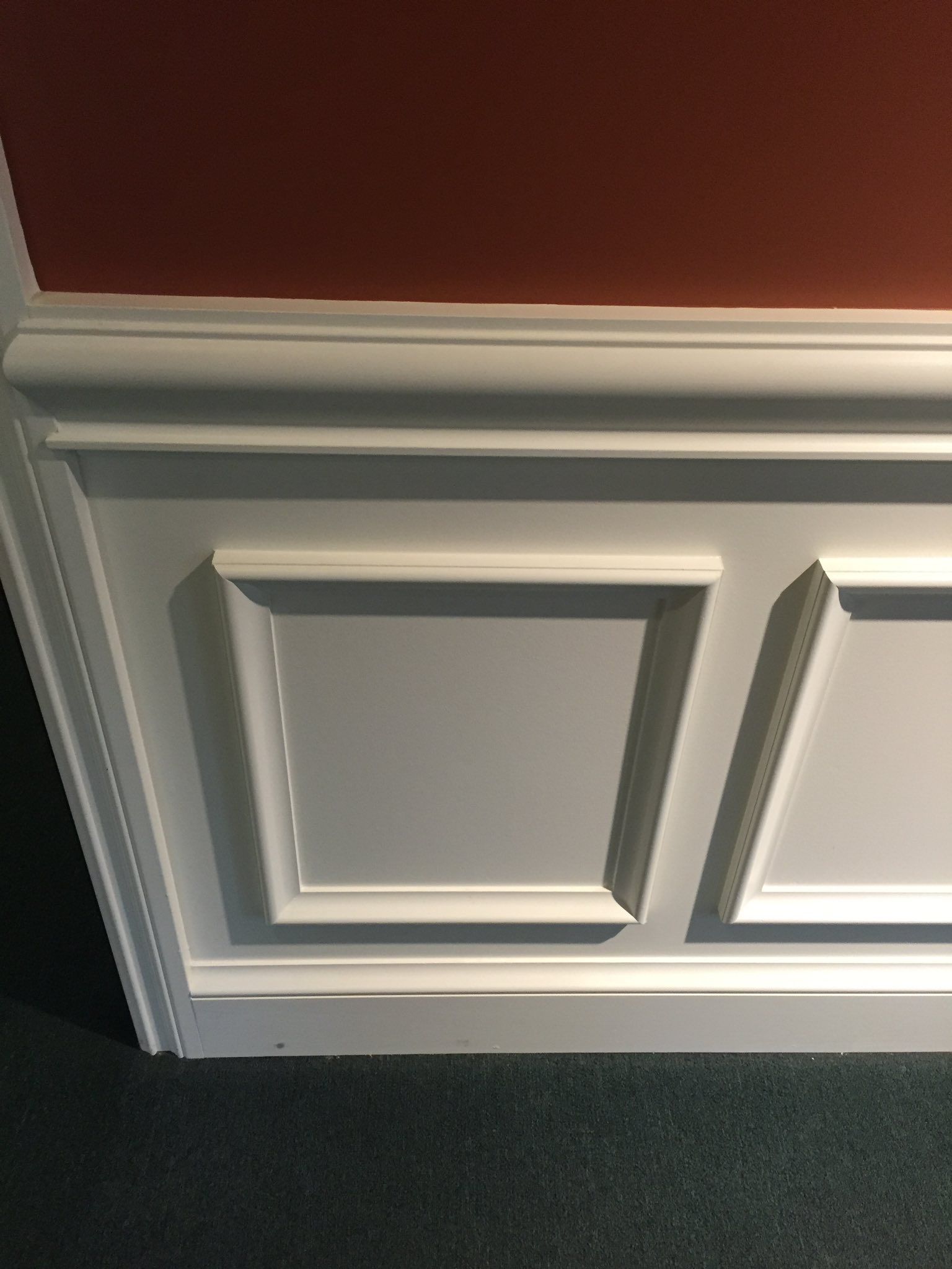 White wainscoting with square panels against a gray wall, below a red upper wall, on green carpet.