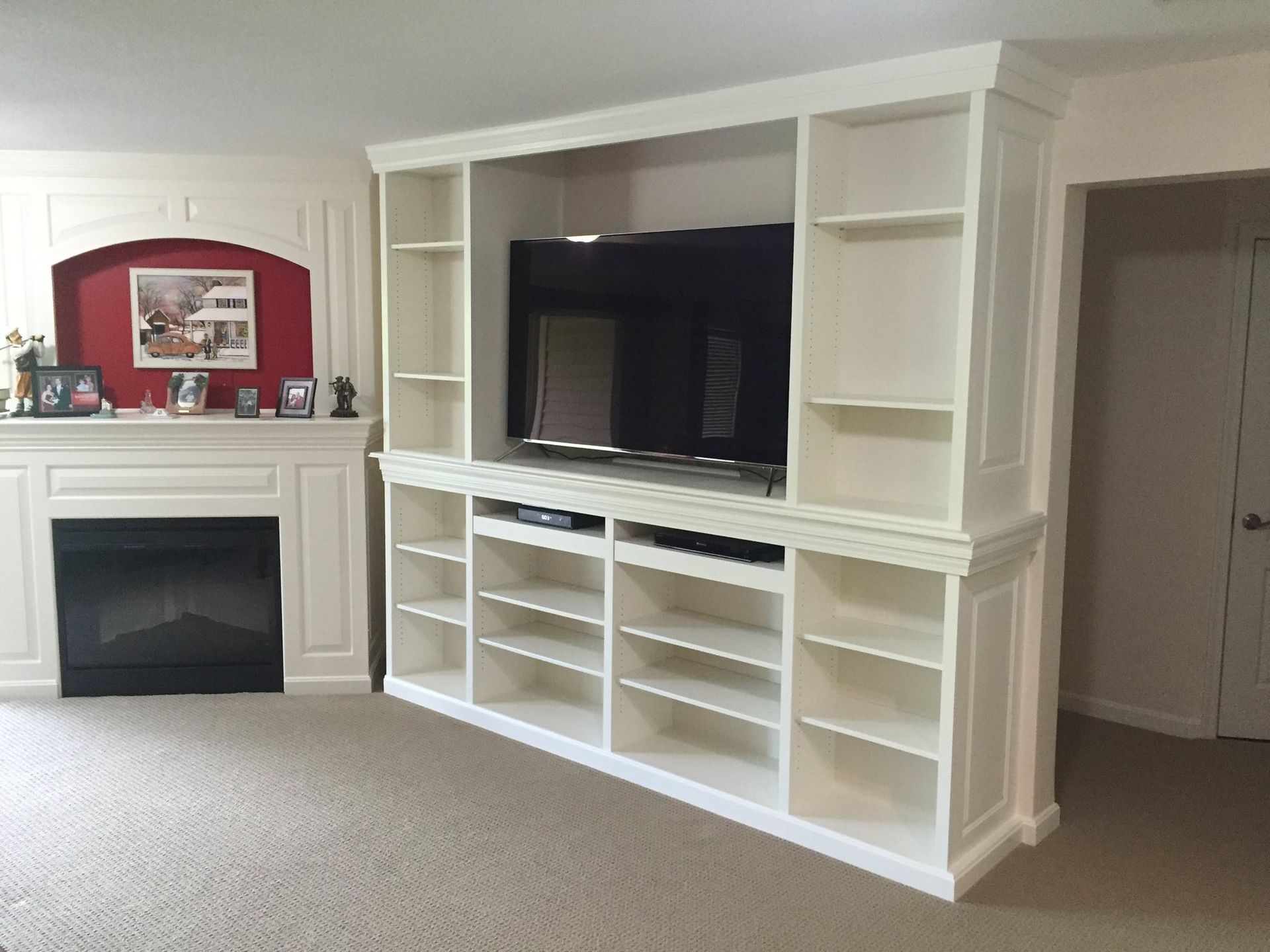 Built-in white entertainment center with TV and shelves, next to a fireplace. Beige carpet and white walls.