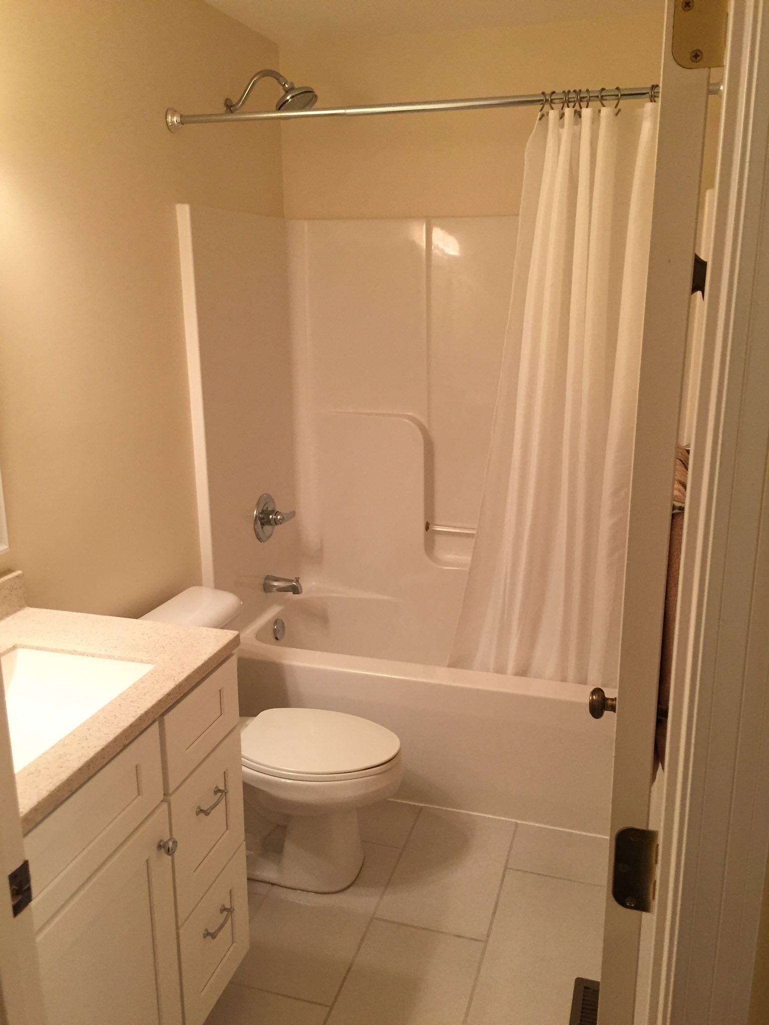 Bathroom with white tub, toilet, and vanity. Beige walls, gray floor tiles, and a white shower curtain.
