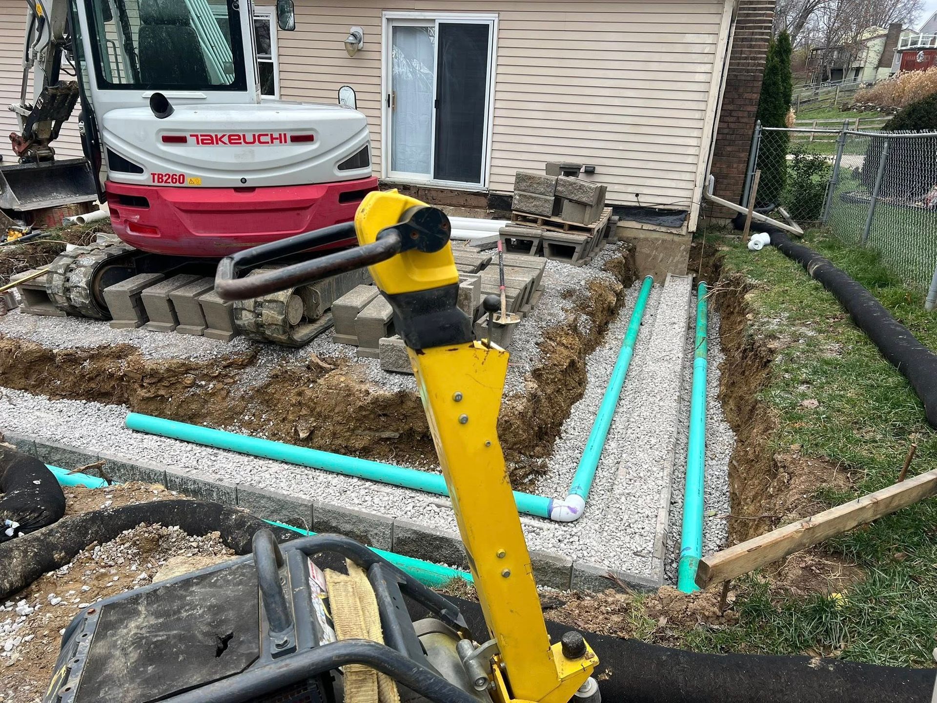 Construction site with excavator and trenches. Green pipes laid in gravel, near a house.