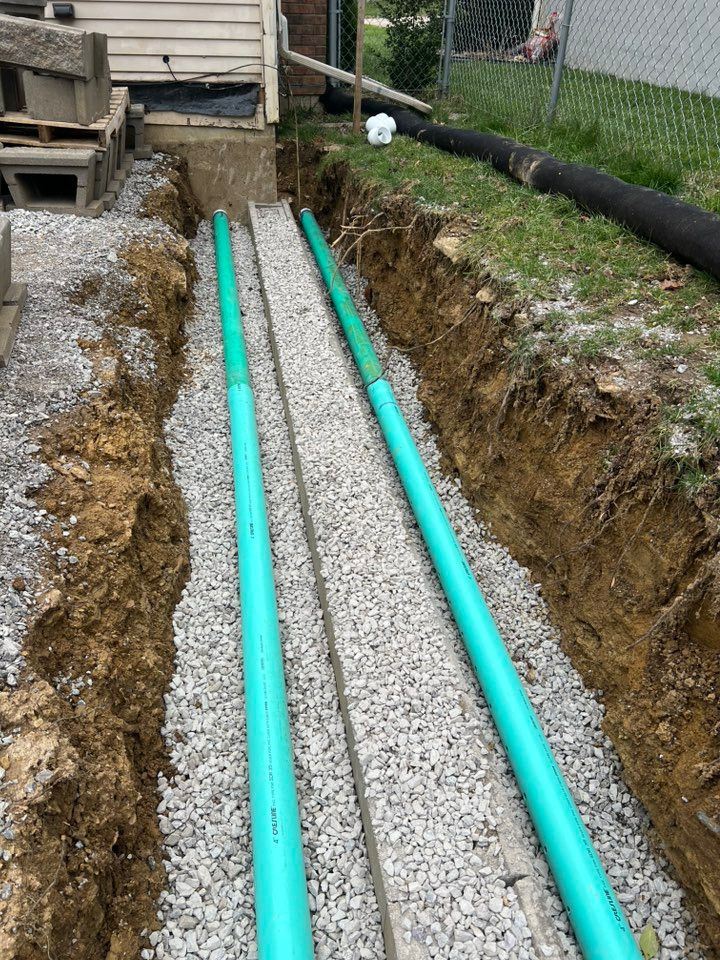 Trenches with green pipes laid in gravel, near a building and grass.