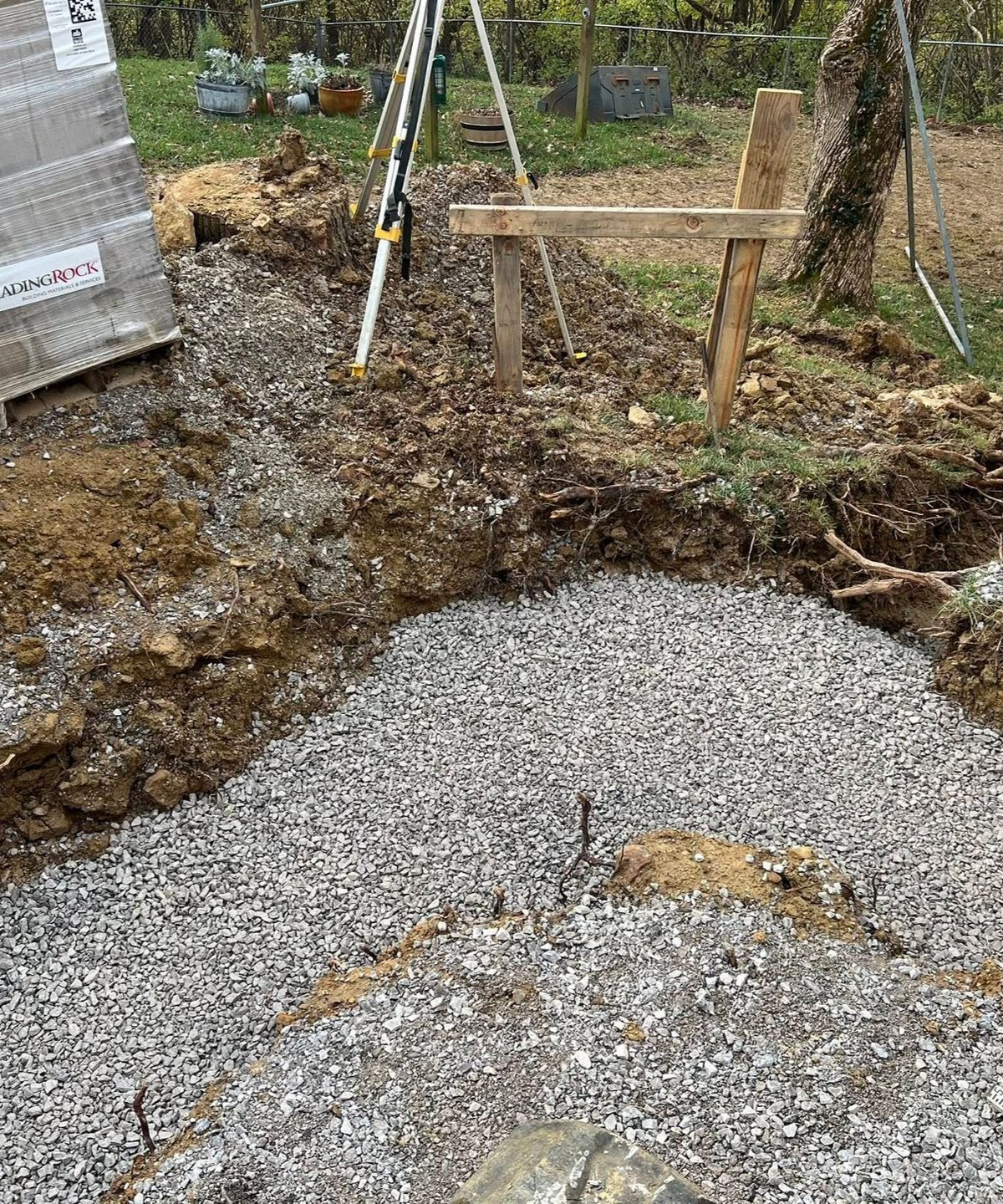 Gravel base in an excavated area, with wooden stakes and a tripod in a yard.