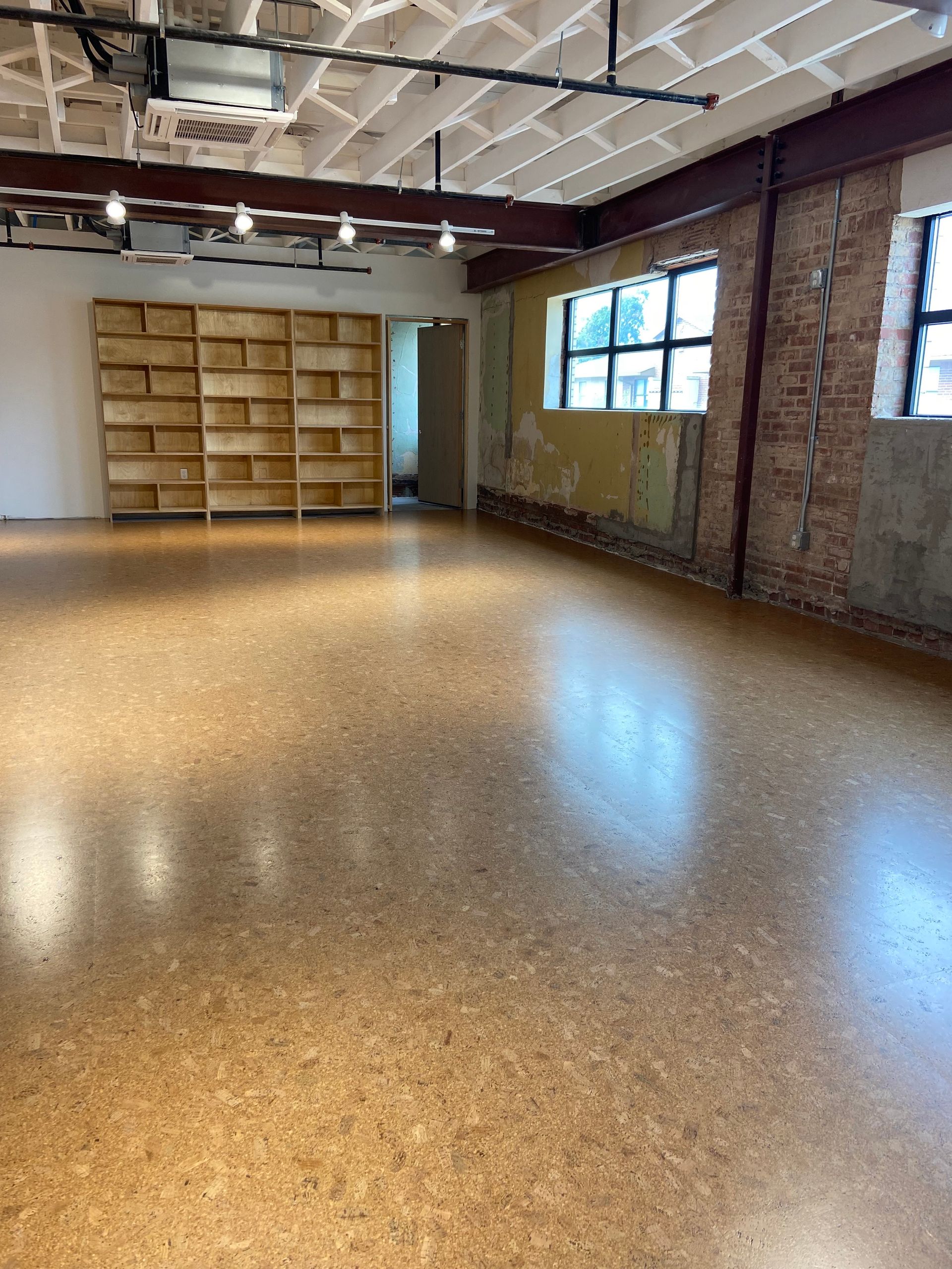 Empty industrial-style room with brick walls, large windows, and shelving along the back wall.