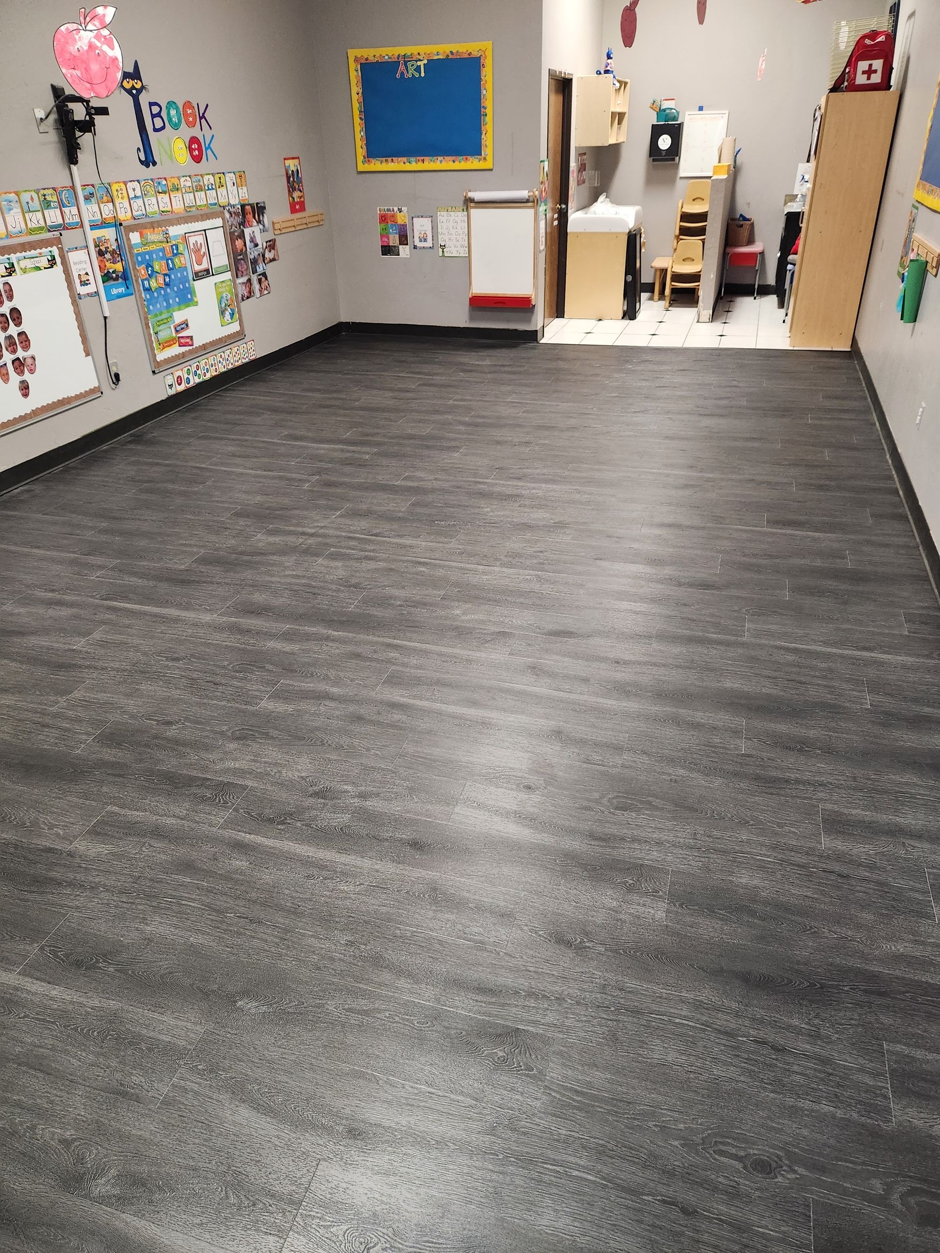Empty classroom with a dark gray floor, white walls, colorful wall art, and a small work area in the back.