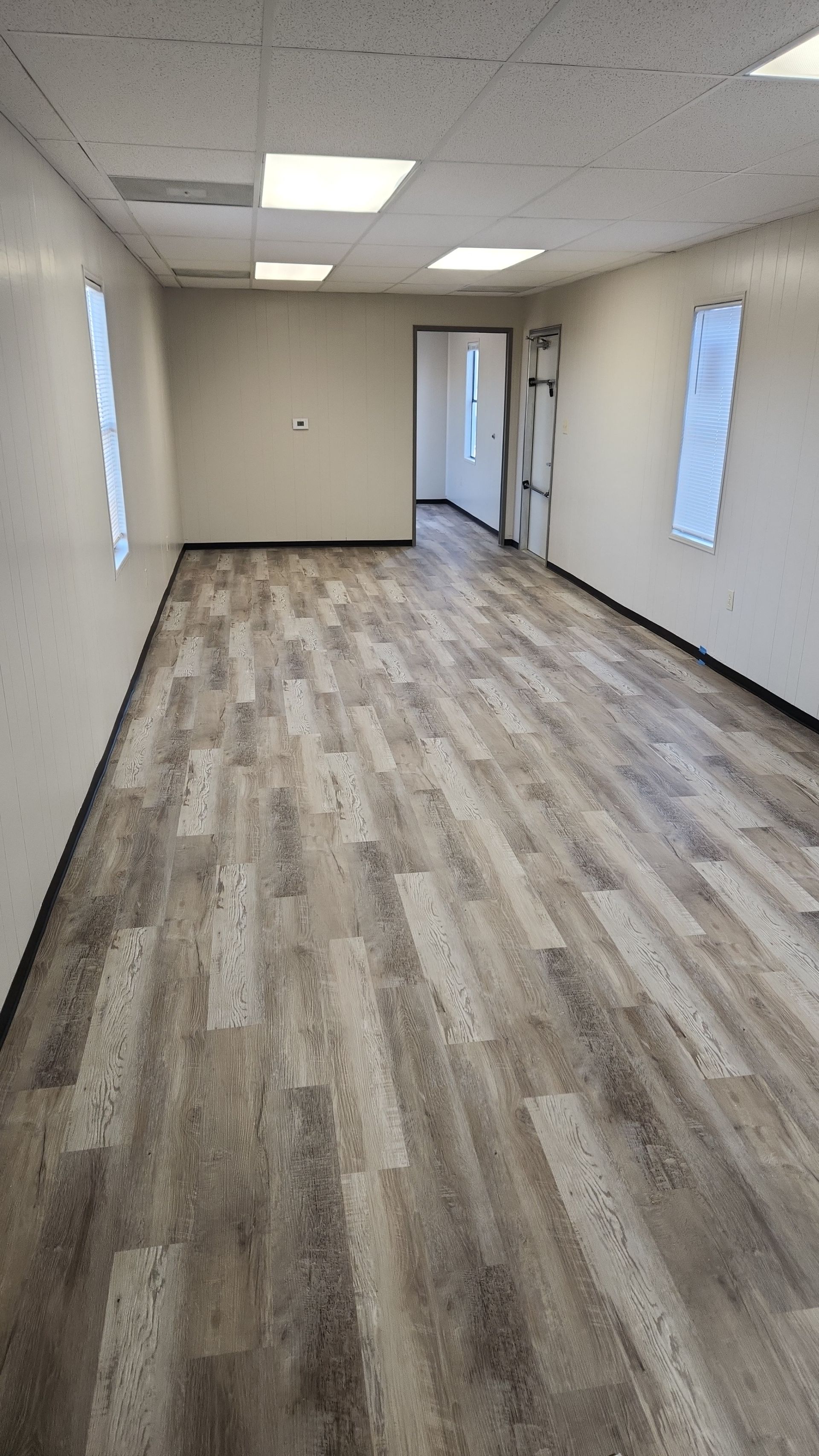 Empty office room with wood-look flooring, white walls, windows, and a drop ceiling with fluorescent lights