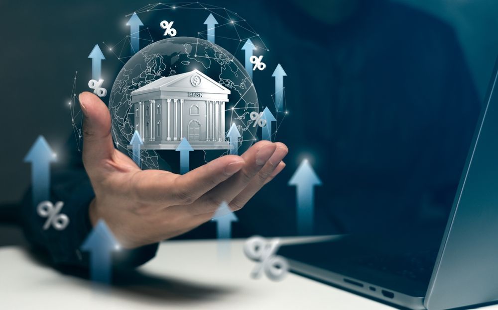 Hand holding a bank icon in a glowing sphere with arrows and percent signs beside a laptop