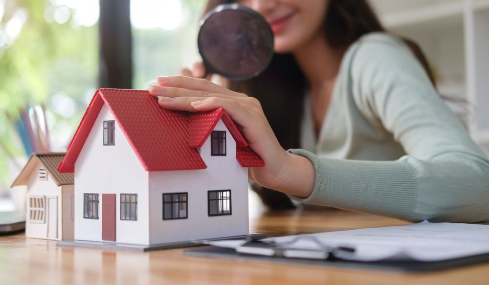 Hands placing a magnifying glass over a model house beside a laptop and notebook