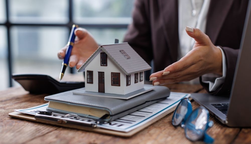 Hands gesturing over a model house on a desk, with a pen, notebook, and laptop in a business setting