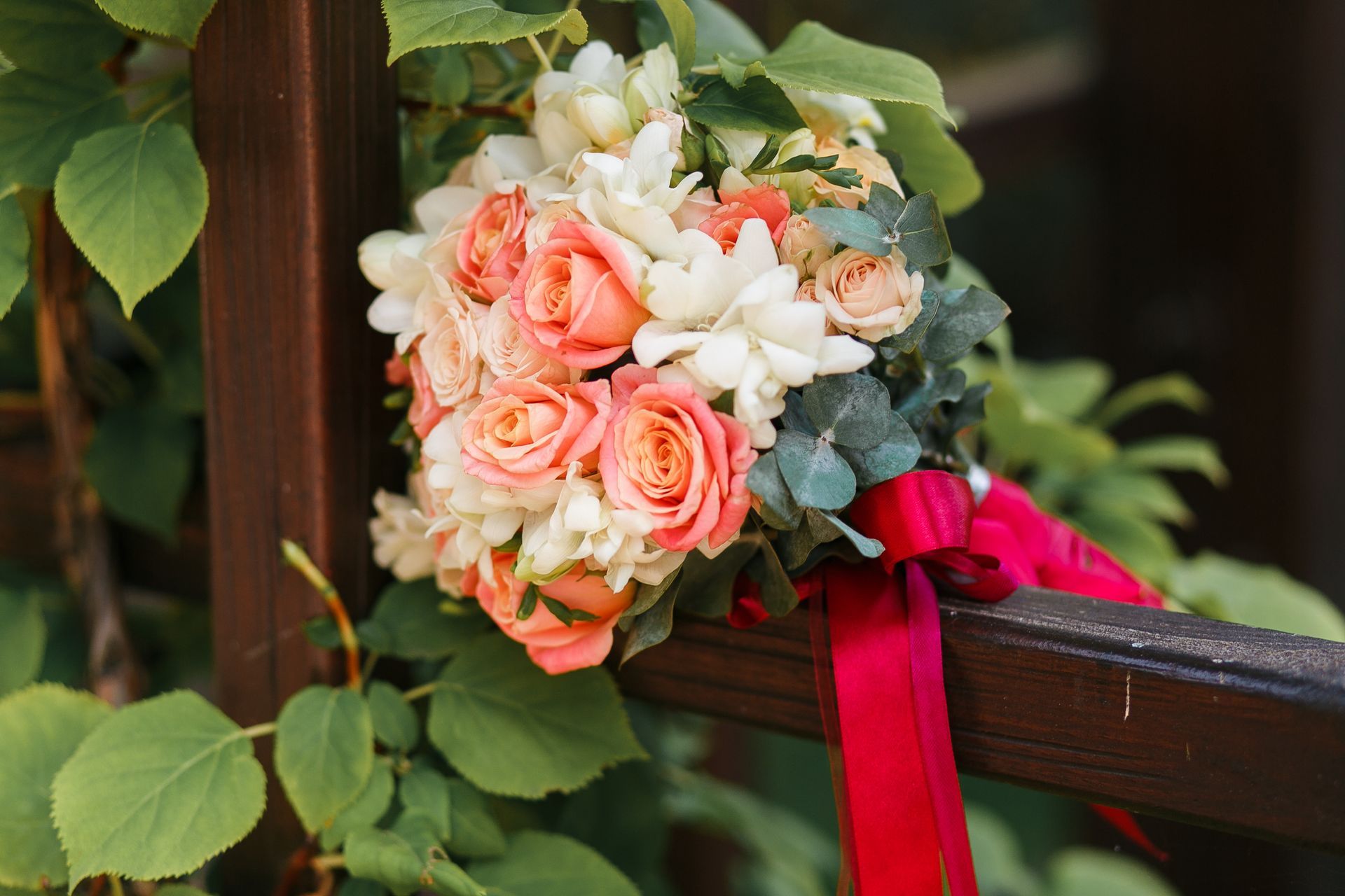 Pink and white rose bouquet with greenery and red ribbon on a wooden railing