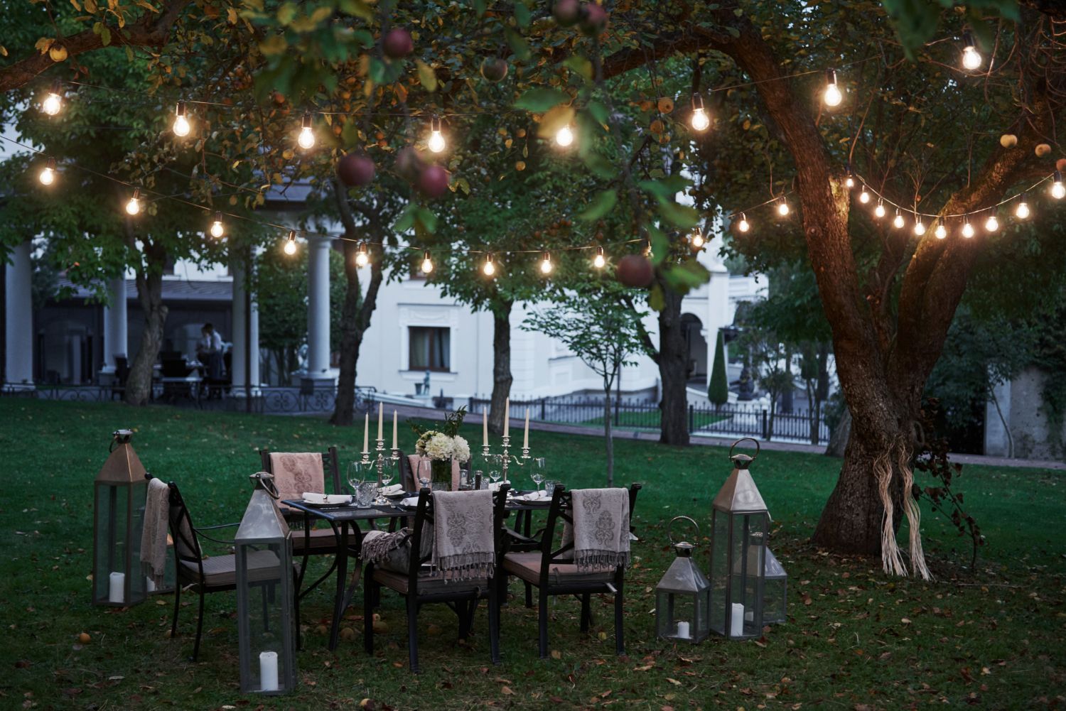 Outdoor dining table set under a tree with string lights, lanterns, and a white building in the background.