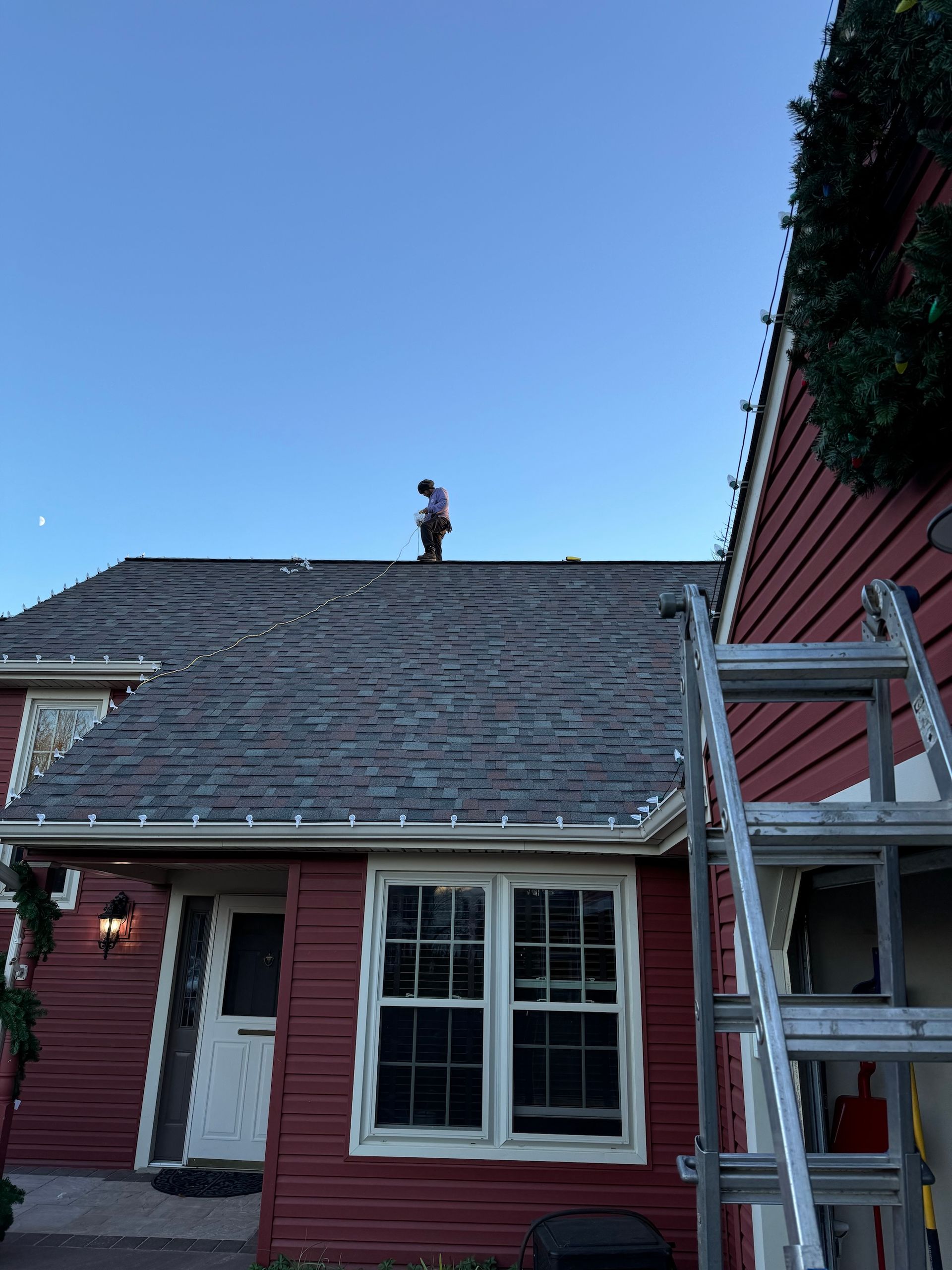 Person on a rooftop installing Christmas lights on a red house with a ladder.