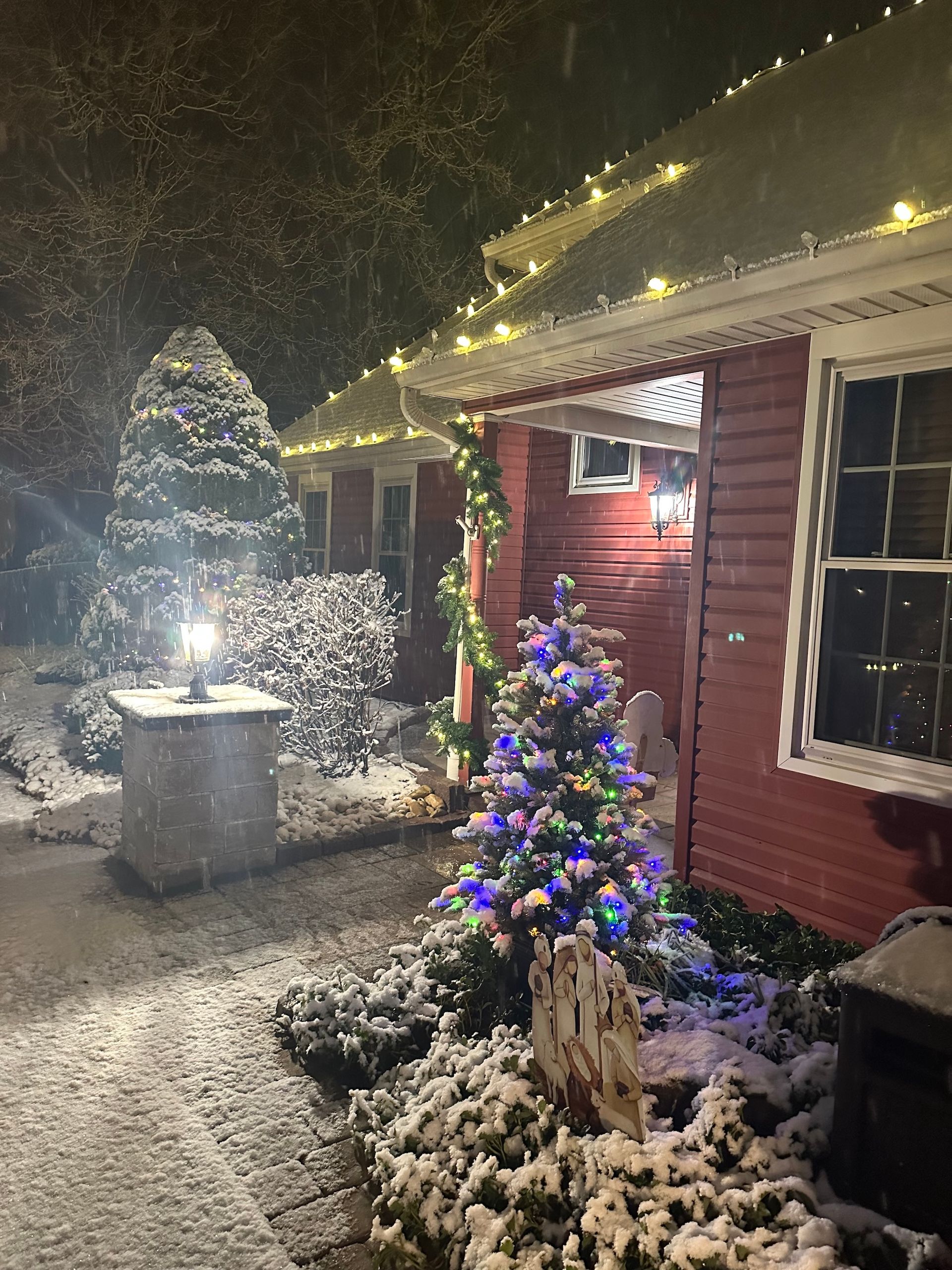 Snowy scene with a red house lit with string lights and a Christmas tree, lantern, and bushes.