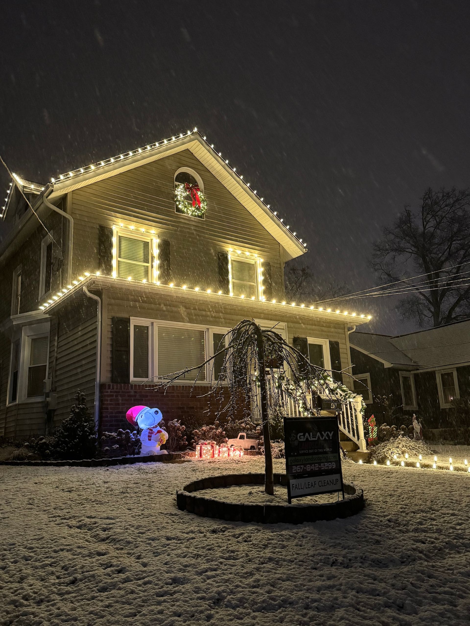 Snowy house with Christmas lights, a Snoopy display, and a wreath.