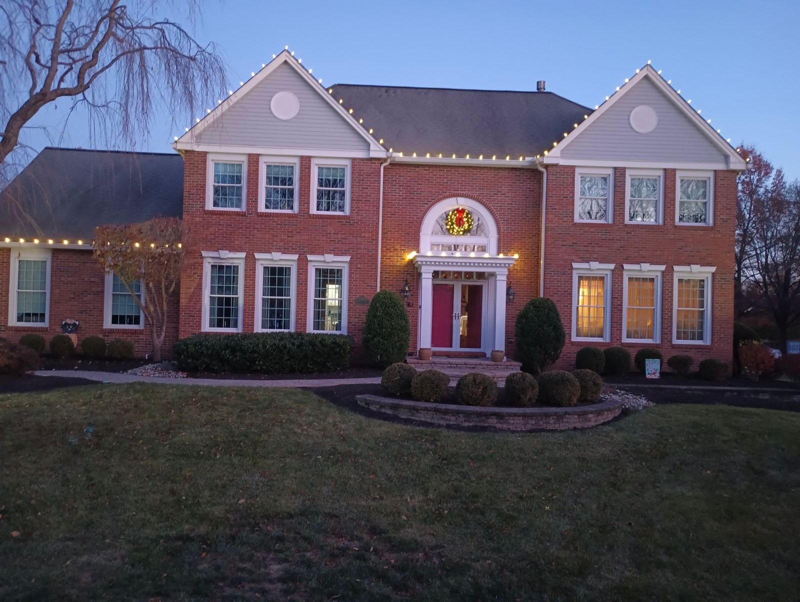 Two-story brick house at dusk, lit with white lights. Christmas wreath over red front door.