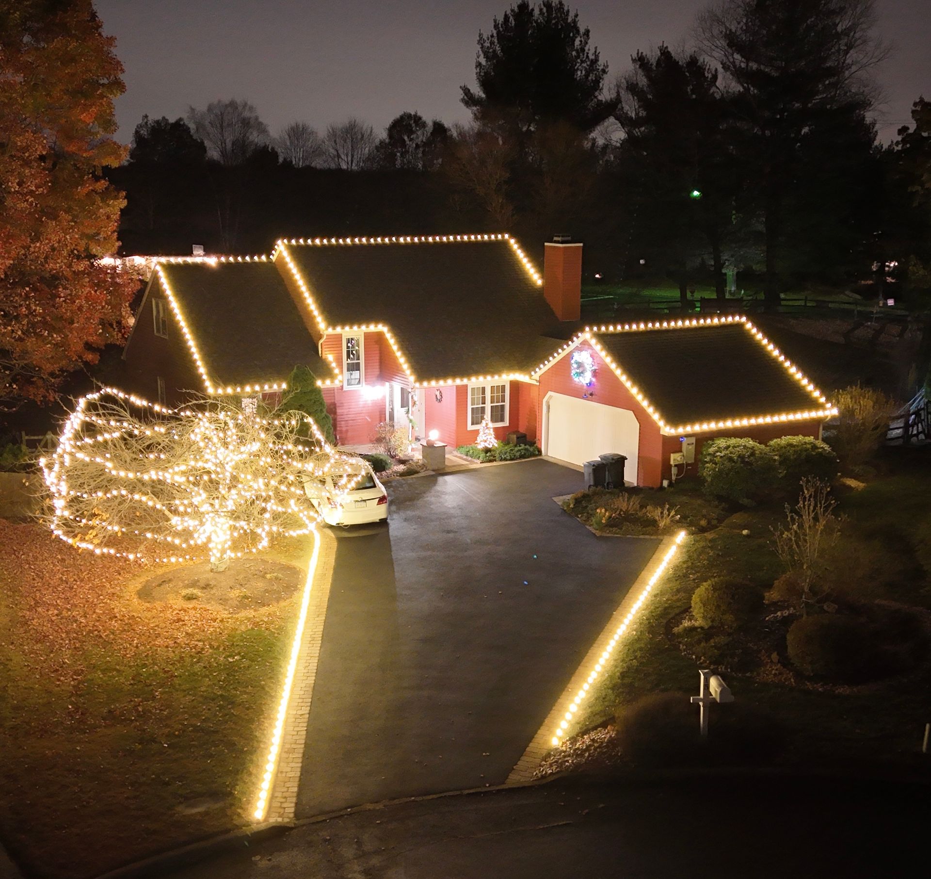 House decorated with yellow Christmas lights, including rooflines, driveway, and a tree, at night.