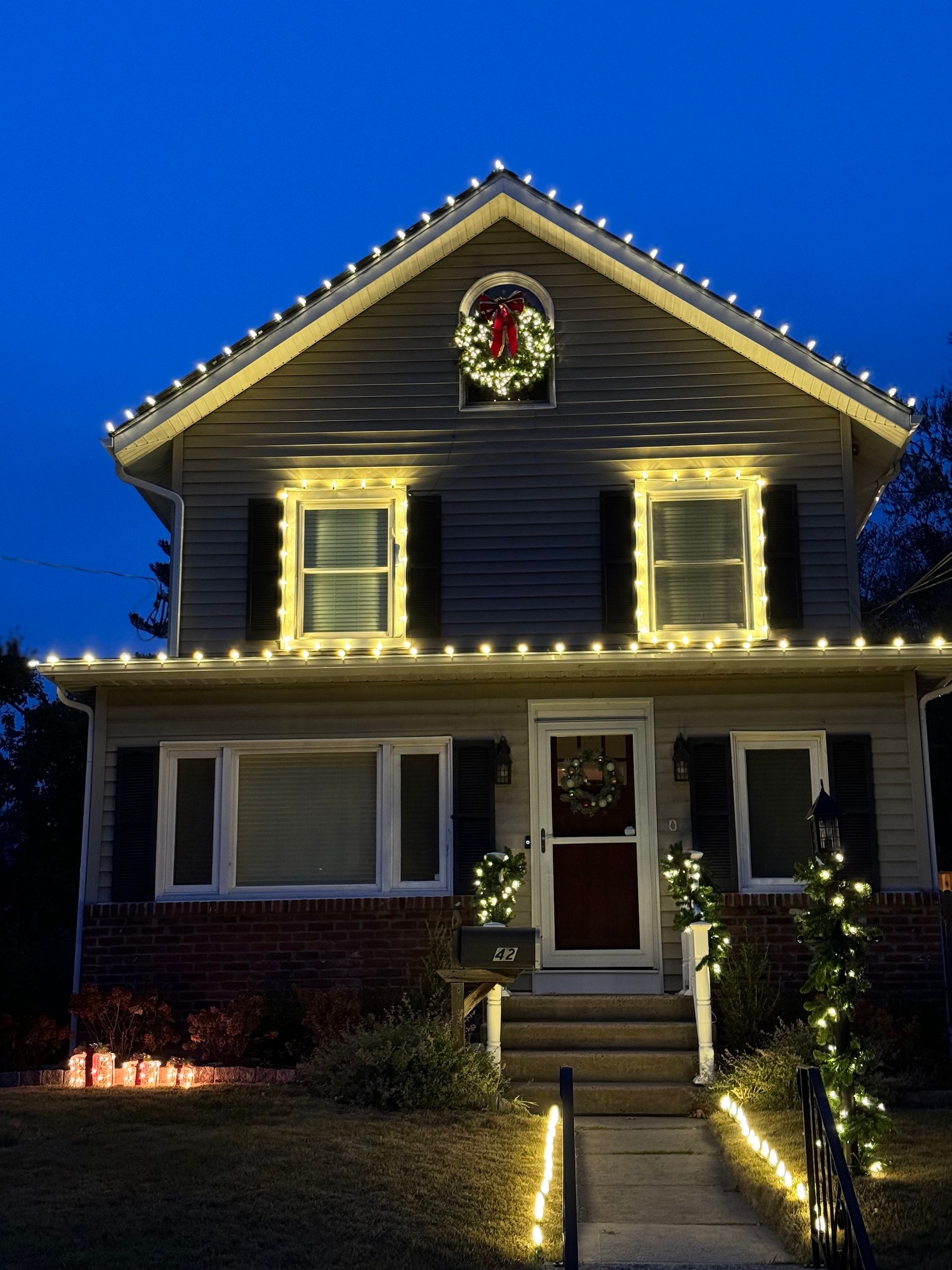 House decorated with Christmas lights at dusk, wreath on the door and window, string lights outlining the roof.