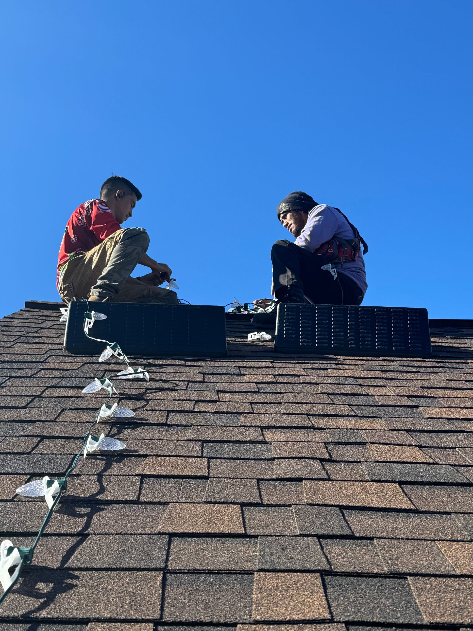 Two people on a brown shingled roof, working under a clear blue sky.