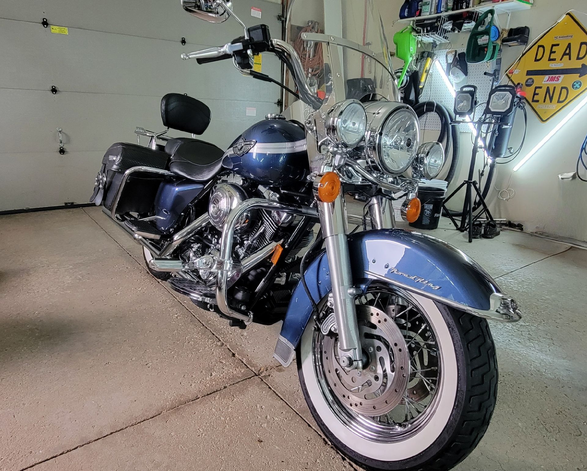 Blue motorcycle in a garage with a whitewall front tire and chrome details, parked beside a wall sign.