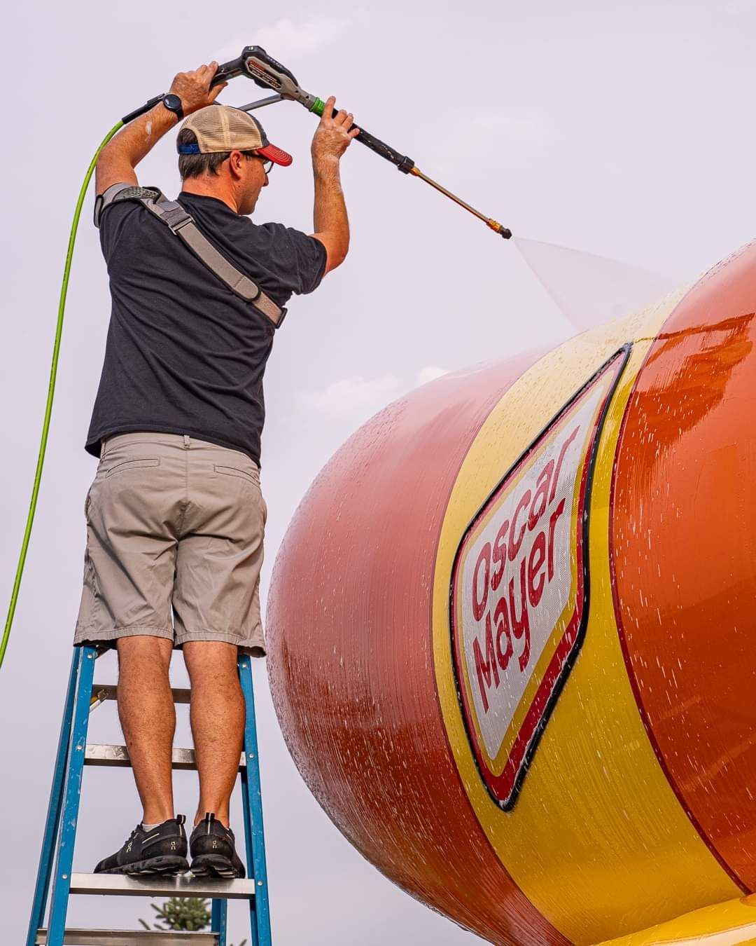 Worker on a ladder pressure-washing a giant orange and yellow buoy or tank with a hose.